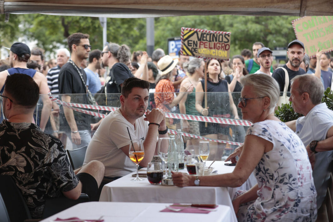 Turistas en un bar de Barcelona observan las protestas en su contra por parte de vecinos molestos. Foto de la agencia EFE