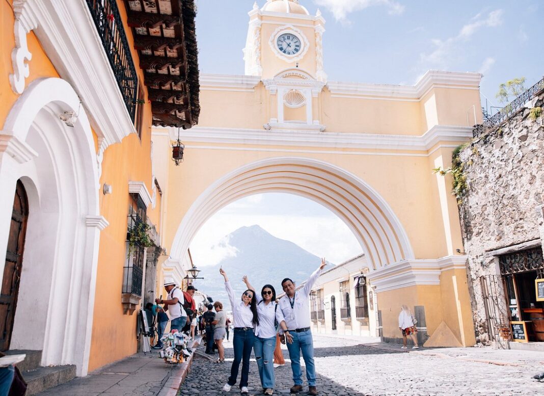 Un grupo de turistas posando frente al icónico Arco de Santa Catalina en Antigua Guatemala, con el majestuoso Volcán de Agua al fondo.